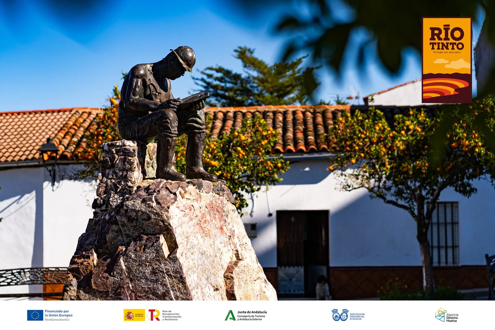 Monumento de bronce a un minero sentado sobre una roca en Campofrío, frente a una casa blanca y unos naranjos, junto a la iglesia de San Miguel Arcángel.