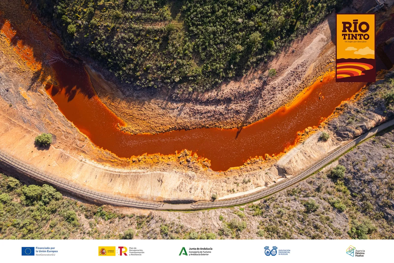 Vista aérea del Río Tinto serpenteando en naranja entre riberas verdes y un camino de tierra que lo acompaña.