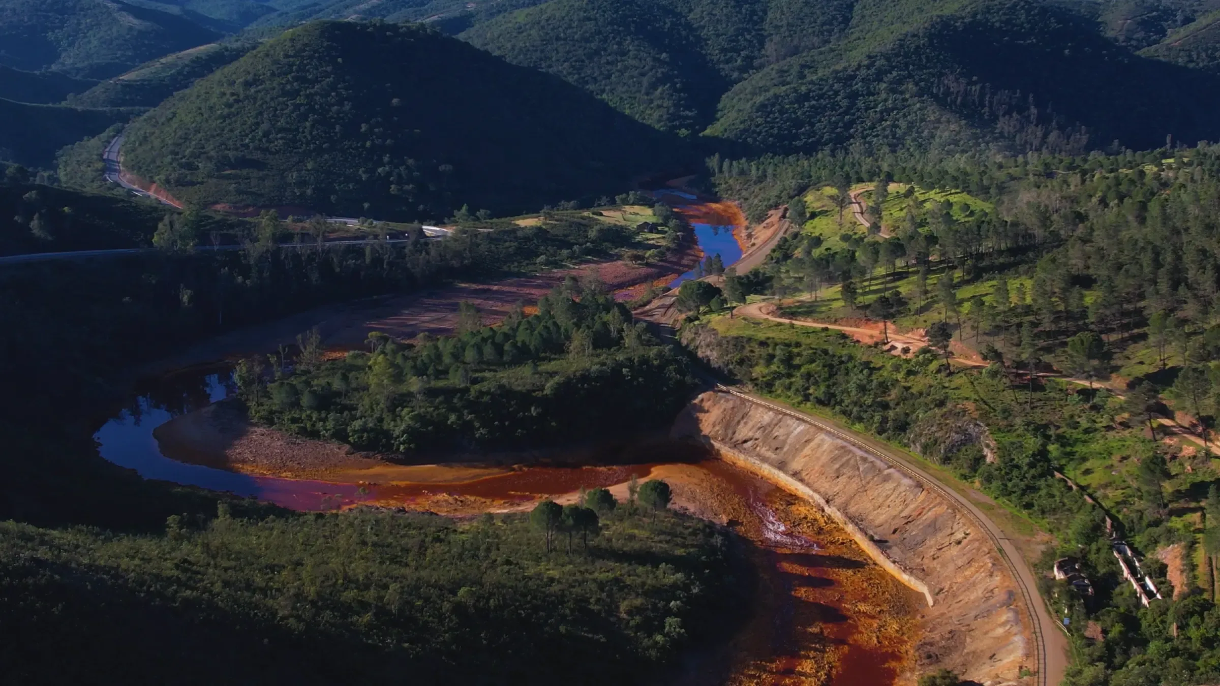 El río Tinto desde el aire: el agua roja traza una curva entre lomas de pinar, con la vía del tren minero en la orilla, en un fotograma de la campaña de Bis para la Cuenca Minera