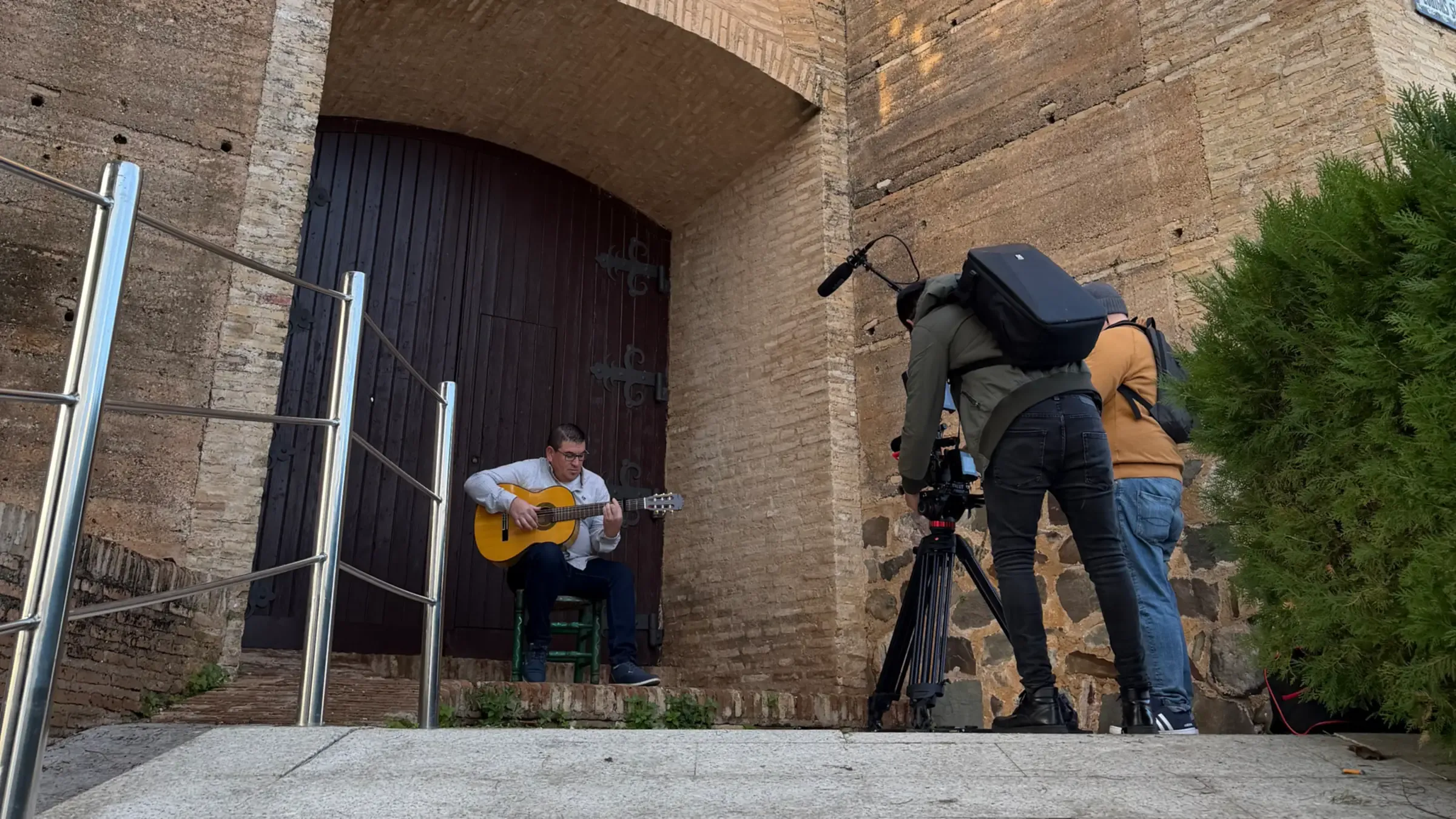 Rodaje de Bis en exteriores: un guitarrista toca sentado en la puerta de un castillo mientras dos personas del equipo graban con cámara sobre trípode y micrófono de caña