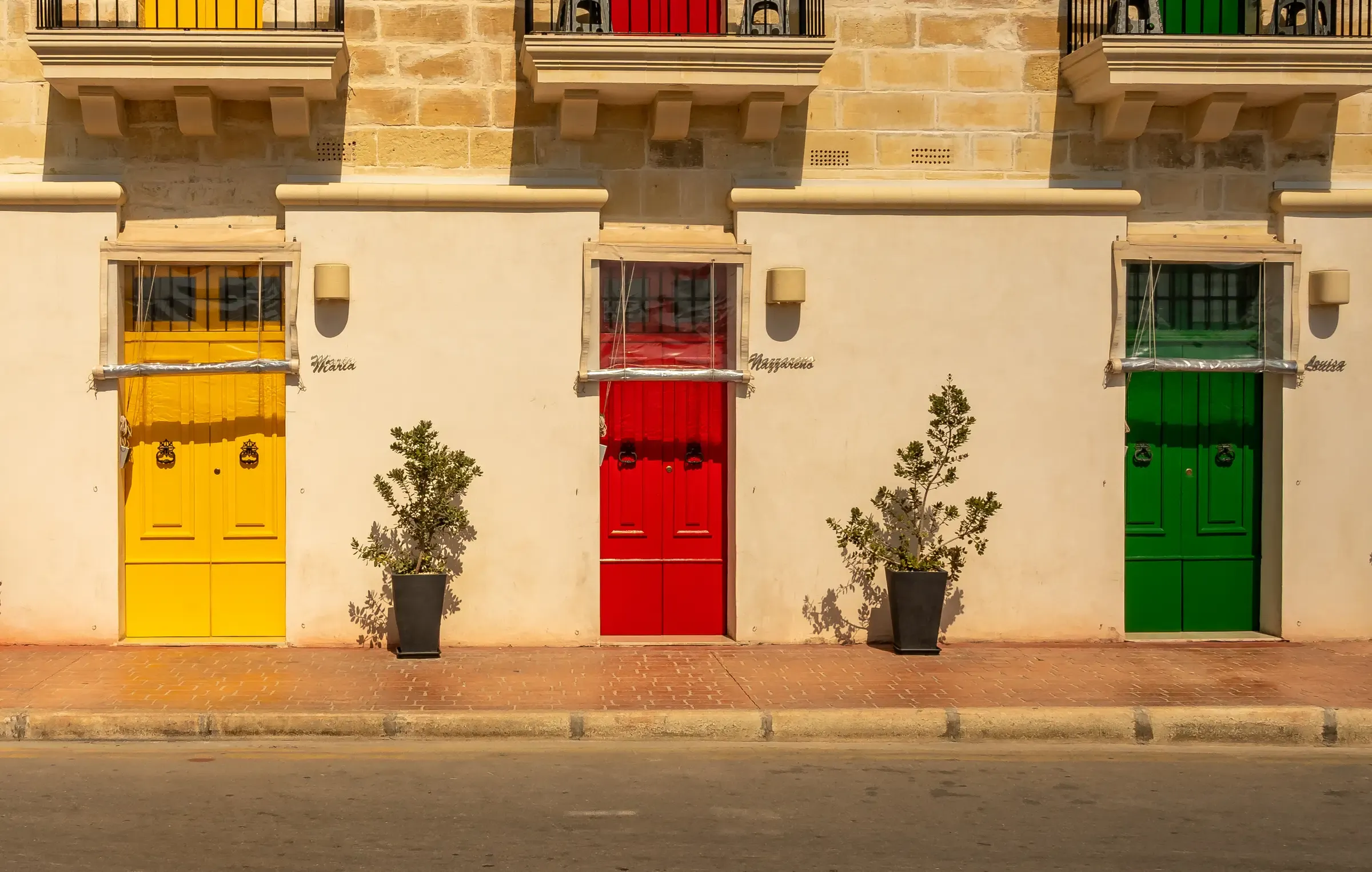 Tres puertas iguales en una misma fachada, pintadas de amarillo, rojo y verde, con una maceta entre cada una